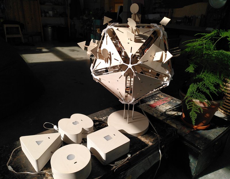 Photo of a wooden virus (20 sided wood shape) with different shapes sticking out of each face, and below it are four different shaped wood boxes each with a tiny black sensor visible and a frosted white window that lights up (the boxes are shaped like a guitar, square, circle and triangle).