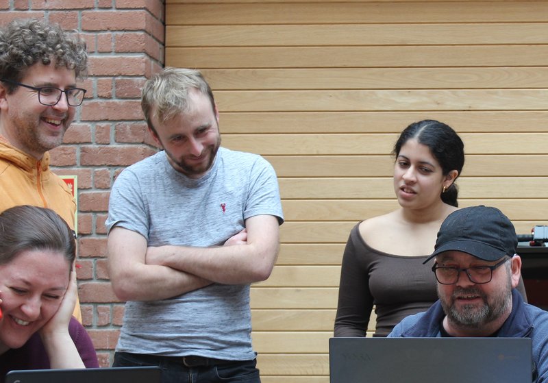 photo of 5 people, two are playing the game at laptops and three are looking over their shoulders, everyone is either laughing or looking quizzical