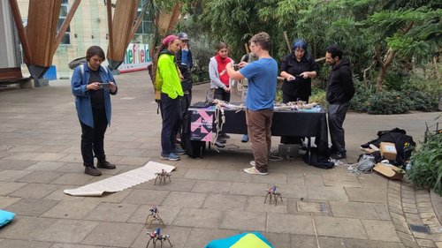 A group of people around a table with plants in the background and robots on the floor.